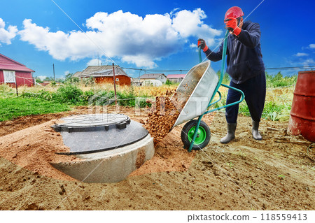 Worker buries concrete rings of septic tank with sand, pouring it out of wheelbarrow. 118559413