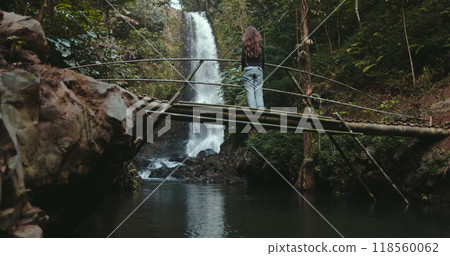Young woman is enjoying the view of a waterfall in a tropical rainforest while standing on a bamboo bridge. She is surrounded by lush greenery and the sound of cascading water 118560062