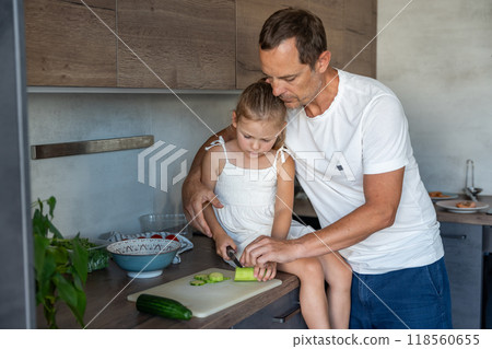 Father patiently teaching a his daughter how to cook in their home kitchen, family with dad and daughter preparing green salad with vegetables  118560655