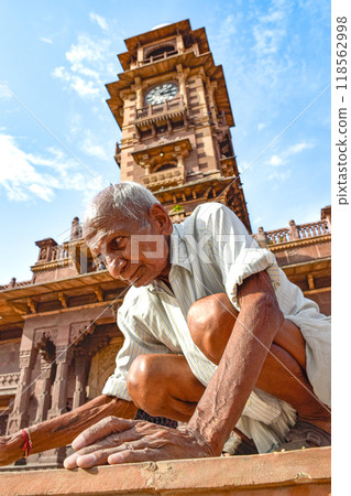 Indian elderly man with white hair under clock square in brown tones crouching 118562998