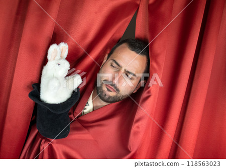 Funny young adult man magician with interesting face playing with rabbit out hat Funny young adult man magician with interesting face playing with rabbit out hat 118563023