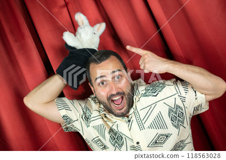 Funny young adult man magician playing with rabbit out hat with red background Funny young adult man magician playing with rabbit out hat with red background 118563028
