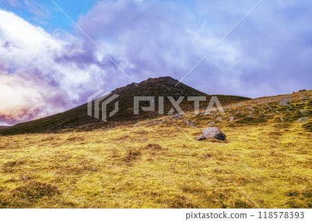 A grassy hillside slopes upward towards a mountain peak under a partly cloudy sky. Scattered rocks dot the foreground of the landscape. A grassy hillside slopes upward towards a mountain peak under a partly cloudy sky. Scattered rocks dot the foreground of the landscape. 118578393