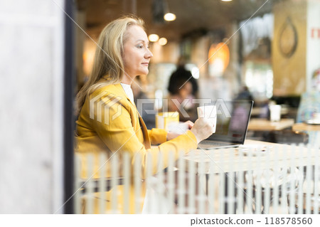 Business lunch - businesswoman sits at table in cafe by window with a laptop and drinks coffee. View through window 118578560