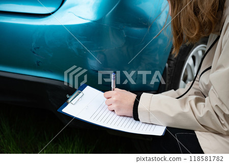 Person inspecting car bumper damage with clipboard outdoors. 118581782