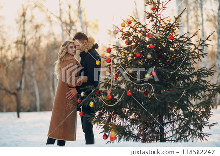 Young adult couple man and woman decorates pine tree in winter park forest. New year tree holiday party celebration concept Young adult couple man and woman decorates pine tree in winter park forest. New year tree holiday party celebration concept 118582247
