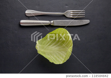 Fresh green cabbage leaf and table knife with fork on a black stone table. Vegetarian raw food diet concept Fresh green cabbage leaf and table knife with fork on a black stone table. Vegetarian raw food diet concept 118584842