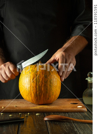 Chef tries to cut ripe pumpkin on cutting board with knife. Fruit diet concept in restaurant kitchen. American national cuisine or making pie 118591591