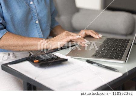 Woman using laptop sit at desk, calculator and documents nearby Woman using laptop sit at desk, calculator and documents nearby 118592352