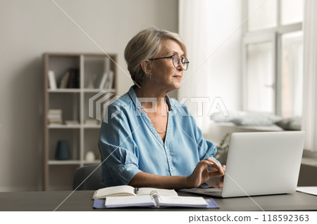 Woman sits at desk with laptop, looking thoughtfully out window Woman sits at desk with laptop, looking thoughtfully out window 118592363