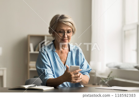 Senior woman sitting at desk using modern smartphone Senior woman sitting at desk using modern smartphone 118592383
