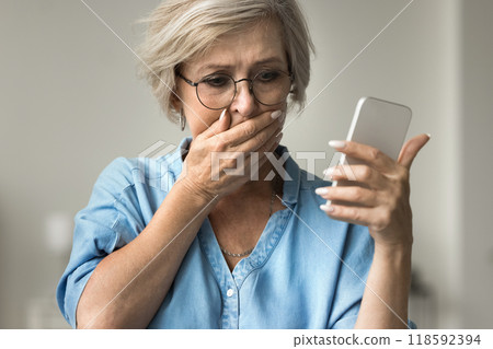 Closeup older woman looks at smartphone screen with distressed expression Closeup older woman looks at smartphone screen with distressed expression 118592394