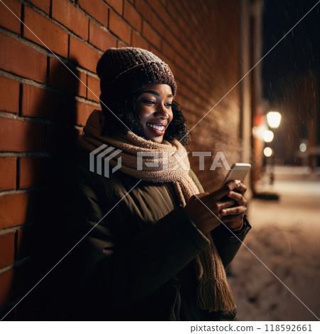 A woman in winter clothes leans against a snow-dusted brick wall, smiling at her phone. Her breath is visible in the chilly air, with cozy street lights creating a warm atmosphere 118592661