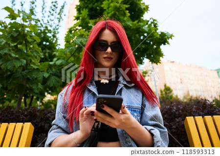 Young woman with red hair uses smartphone while sitting on a park bench Young woman with red hair uses smartphone while sitting on a park bench 118593313