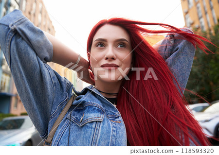 Young woman with vibrant red hair smiles on urban street in sunshine 118593320