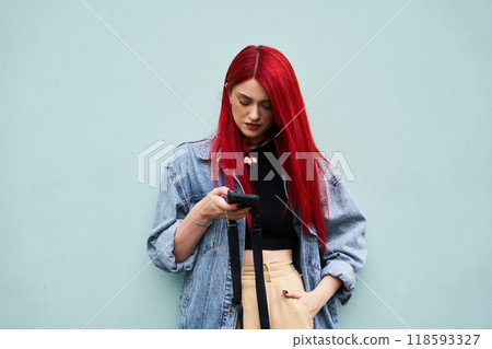 Young woman with red hair checks her phone against a light blue wall 118593327