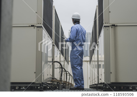 Building maintenance image: A man in work clothes inspecting the outdoor air conditioning unit installed on the roof of a building 118593506