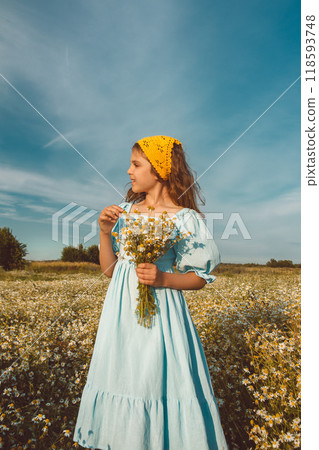 Happy natural girl in blue linen dress and yellow knitted kerchief stands and holds bouquet of flowers in daisy field Happy natural girl in blue linen dress and yellow knitted kerchief stands and holds bouquet of flowers in daisy field 118593748