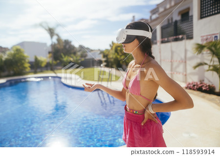 A woman wearing a virtual reality headset while relaxing by the poolside in a sunny paradise A woman wearing a virtual reality headset while relaxing by the poolside in a sunny paradise 118593914