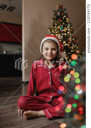 Happy girl of 7 years old in red pajamas and Santa Claus hat sitting on living room floor next to Christmas tree 118594378