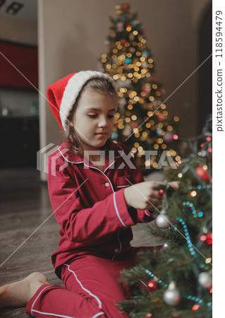 Portrait of girl of 7 years old in red pajamas and Santa Claus hat sitting on living room floor decorates Christmas tree Portrait of girl of 7 years old in red pajamas and Santa Claus hat sitting on living room floor decorates Christmas tree 118594479