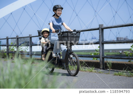 Mom and child riding bicycles on a beautiful road with blue sky. Going out or going to work on a mamachari. Wide-angle shot of the whole body. Greenery is blurred in front. 118594793