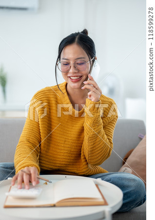 A cheerful Asian woman is sitting on a sofa, talking on the phone while making a list in a notebook. 118599278