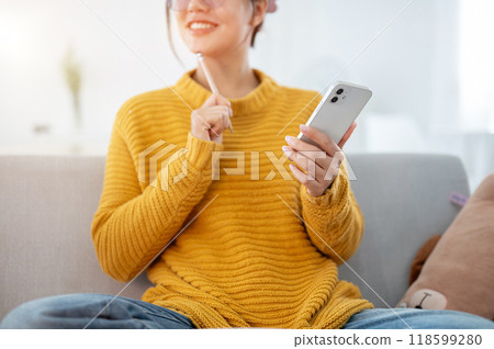 A positive, smiling Asian woman pondering ideas while using her smartphone on a couch at home. 118599280