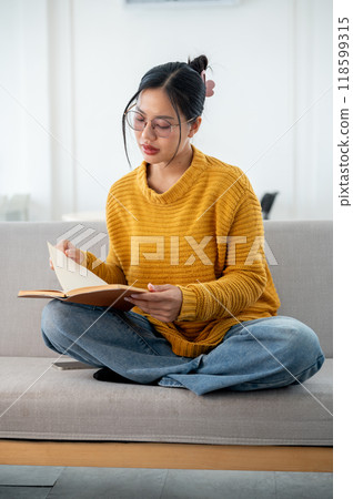 A beautiful Asian woman in casual clothes focuses on reading a book on a couch at home. 118599315