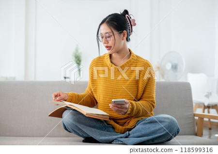 A beautiful Asian woman in casual clothes focuses on reading a book on a couch at home. A beautiful Asian woman in casual clothes focuses on reading a book on a couch at home. 118599316