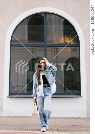 Young woman in a blue jacket and jeans is standing in front of a building. She is smiling and holding her hand up 118601544