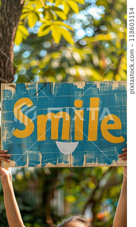 In a vibrant park filled with greenery, an individual shares an uplifting message of happiness by holding a colorful sign that says Smile, symbolizing optimism and joy 118603154