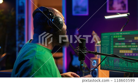 Confident artist singer performing a song on microphone in home studio, recording his audio files and manipulating raw sound on professional editing software. Music production. Camera A. 118603599