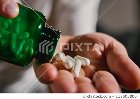 Close-up of man pouring white capsules from green bottle into hand. Person is taking pills or vitamins. Concept of supplements, medicine or health care Close-up of man pouring white capsules from green bottle into hand. Person is taking pills or vitamins. Concept of supplements, medicine or health care 118603806