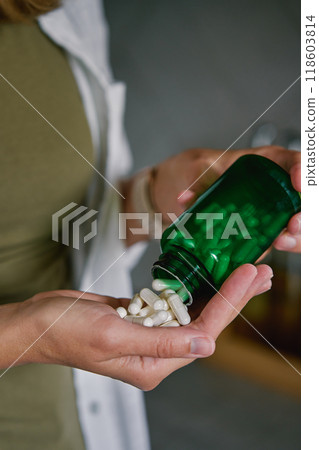 Close-up of woman pouring white capsules from green bottle into hand. Person is taking pills or vitamins. Concept of supplements, medicine or health care 118603814