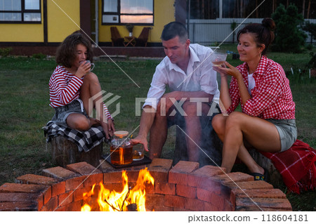 Family mom, dad, daughter are drinking tea in the courtyard of the house by the fire pit on a cozy summer evening. Family mom, dad, daughter are drinking tea in the courtyard of the house by the fire pit on a cozy summer evening. 118604181