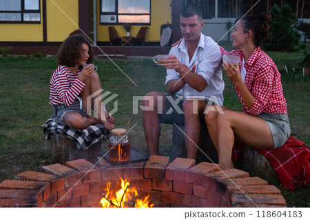 Family mom, dad, daughter are drinking tea in the courtyard of the house by the fire pit on a cozy summer evening. Family mom, dad, daughter are drinking tea in the courtyard of the house by the fire pit on a cozy summer evening. 118604183