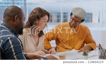 Professional clients and their real estate agent evaluating blueprints for the high class modern space, discussing leasing contract terms before signing. Startup business development. Camera B. Professional clients and their real estate agent evaluating blueprints for the high class modern space, discussing leasing contract terms before signing. Startup business development. Camera B. 118604224