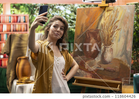 In a modern atelier, artist is filming her rustic artwork painting with the smartphone, social media branding. Recording and taking pictures of colorful canvas with watercolor piece, self promotion. 118604426