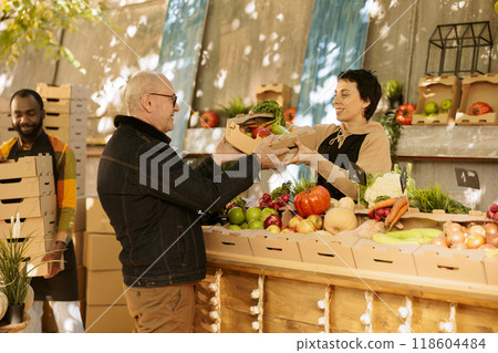 Elderly male customer buying organic natural farm products at farmers market stand, seller giving box of seasonal fresh products. Senior guy carrying colorful farming eco fruits and veggies. 118604484