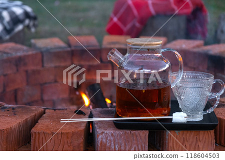 Transparent teapot with tea, cups on a tray on a brick fire pit in the courtyard of the house in a cozy summer evening. 118604503