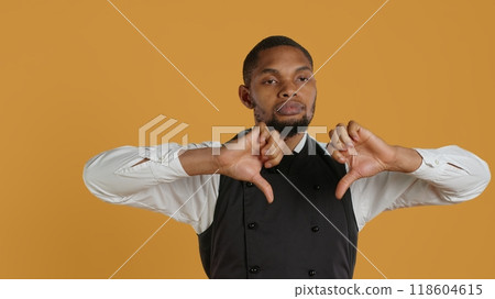 Serious displeased waiter gives a thumbs down sign in studio, presenting his disagreement with a dislike symbol. Disappointed catering employee is dissatisfied, negative gesture. Camera A. 118604615