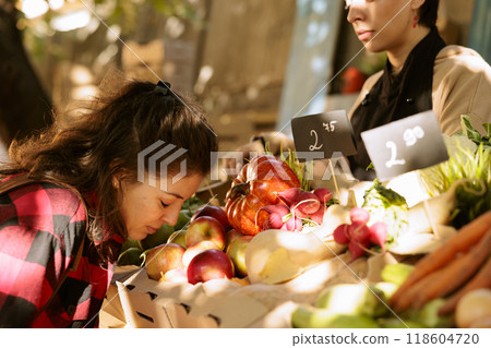 Caucasian female customer smelling freshly harvested red apples from the farmer at agricultural harvest fair booth. Young woman browsing locally grown bio produce on greenmarket stand. Caucasian female customer smelling freshly harvested red apples from the farmer at agricultural harvest fair booth. Young woman browsing locally grown bio produce on greenmarket stand. 118604720
