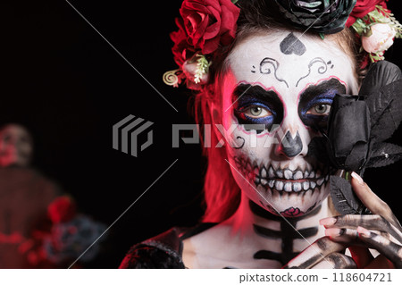 Headshot portrait of woman holding black rose set against dark background celebrating Dia de los Muertos holiday. Close up of lady with flower during Halloween horror event Headshot portrait of woman holding black rose set against dark background celebrating Dia de los Muertos holiday. Close up of lady with flower during Halloween horror event 118604721