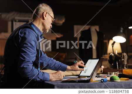 Elderly skilled dressmaker zooming on tablet, looking at sketch drawings of sartorial custom made outfits. Fashion designer working on customer comission in professional atelier shop workspace Elderly skilled dressmaker zooming on tablet, looking at sketch drawings of sartorial custom made outfits. Fashion designer working on customer comission in professional atelier shop workspace 118604738
