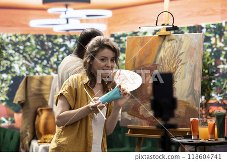 Female artist explaining the painting techniques used on online tutorial, recording a vlog for art class to teach people how to paint with watercolor on canvas. Team of painters do content creation. Female artist explaining the painting techniques used on online tutorial, recording a vlog for art class to teach people how to paint with watercolor on canvas. Team of painters do content creation. 118604741