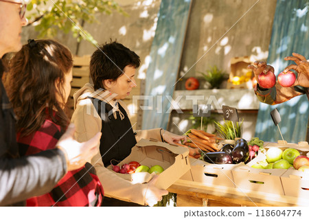 Female merchant selling natural fruits and veggies in boxes, serving senior man and young woman at farmers market. Young vendor picking various freshly harvested produce for customers. Female merchant selling natural fruits and veggies in boxes, serving senior man and young woman at farmers market. Young vendor picking various freshly harvested produce for customers. 118604774
