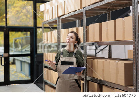 Storage room worker searching clients parcel in storehouse, analyzing clipboard with online order. Warehouse manager wearing uniform overalls supervising merchandise in factory storehouse 118604779