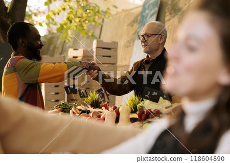 Selective focus of elderly vendor greeting african american client at greenmarket booth, offering fresh organic produce. Smiling multicultural individuals handshaking at farm food market stand. Selective focus of elderly vendor greeting african american client at greenmarket booth, offering fresh organic produce. Smiling multicultural individuals handshaking at farm food market stand. 118604890