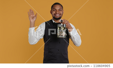 Happy friendly waiter waving hello and welcoming clients at table, holding a cruet set with condiments ready to serve customers. Cheerful lovely staff greeting people at restaurant. Camera B. 118604905